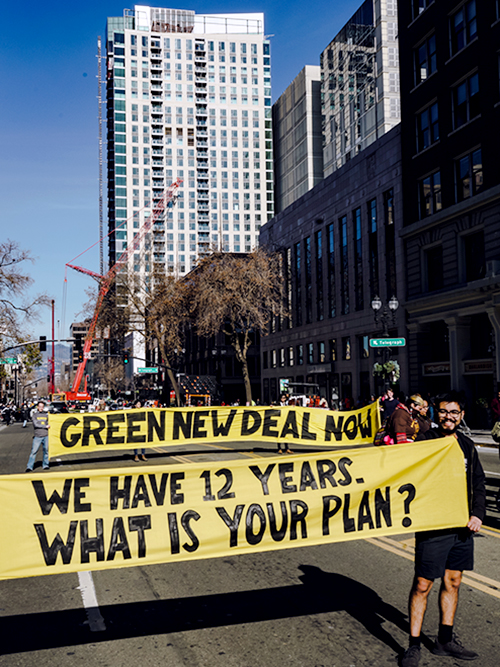 Protesters hold banner signs that stretch across a city road: "GREEN NEW DEAL NOW" and "WE HAVE 12 YEARS. WHAT IS YOUR PLAN?"
