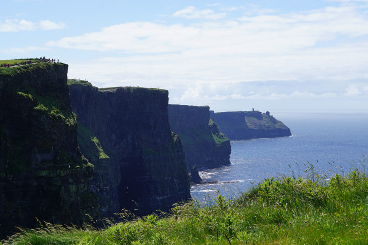 On a sunny day, steep, green-topped cliffs form a wave pattern against blue ocean