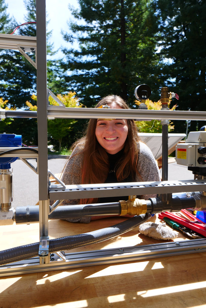 A smiling woman leans against a test station table, with redwoods behind her.