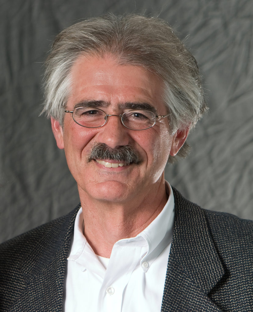 A man with light skin and grey hair, glasses, a white shirt, and a tweed coat, smiles while standing against a grey photo curtain.