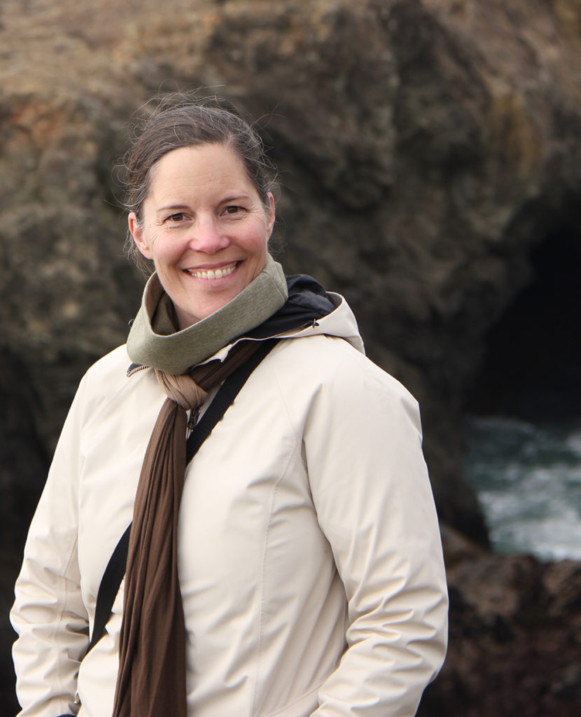 A smiling woman in an off-white coat and scarves stands before a rock face at the ocean.