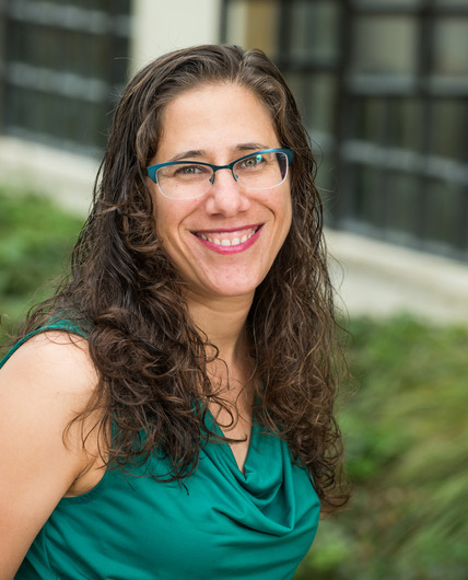 A smiling woman with long hair and glasses is wearing a green shirt and standing outside a building.