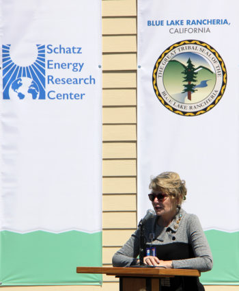 A woman standing in full sun speaks into a podium microphone, with logo banners for the Schatz Energy Research Center and the Blue Lake Rancheria hanging on the wall behind her.