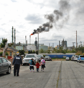 A family walks along a residential street, at the end of which is a refinery with a smoky plume
