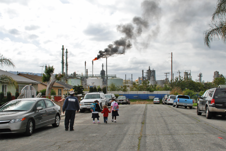 A family walks along a residential street, at the end of which is a refinery with a smoky plume