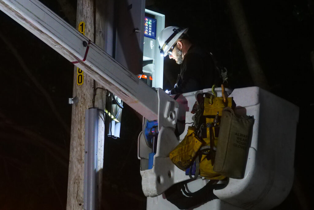 A PG&E technician in a bucket crane works at an illuminated open circuit panel