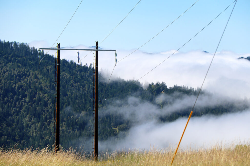 Wires run between transmission poles, with tree-covered hills and clouds in the distance