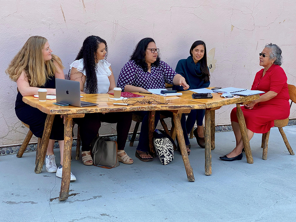 Five women of diverse backgrounds and ages hold a meeting at a work table outside