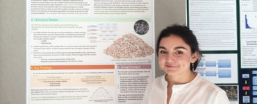 Cassidy Barrientos stands in front of the project poster, wearing a white blouse and a nametag