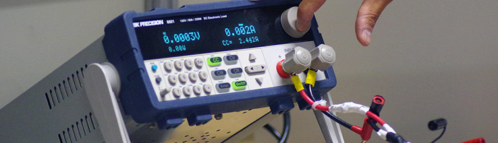 An extended hand turns the dial on an Electronic Load, which reads 0.0003V and 0.002A.