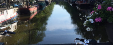 A bike is propped against a bar on a canal bridge. Boats are in the water, flowers and bushes are on the edges, and cars are parked above the canal.