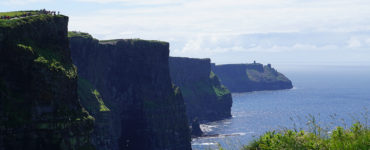 On a sunny day, steep, green-topped cliffs form a wave pattern against blue ocean