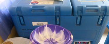 A blue, top-loading refrigerator with a manual on top sits behind nested purple and white plastic bowls and a tan plastic pitcher