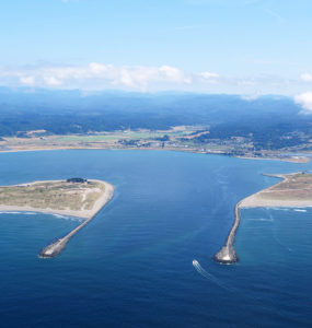 An aerial view of the entrance to Humboldt Bay, where a small boat enters between two jetties.