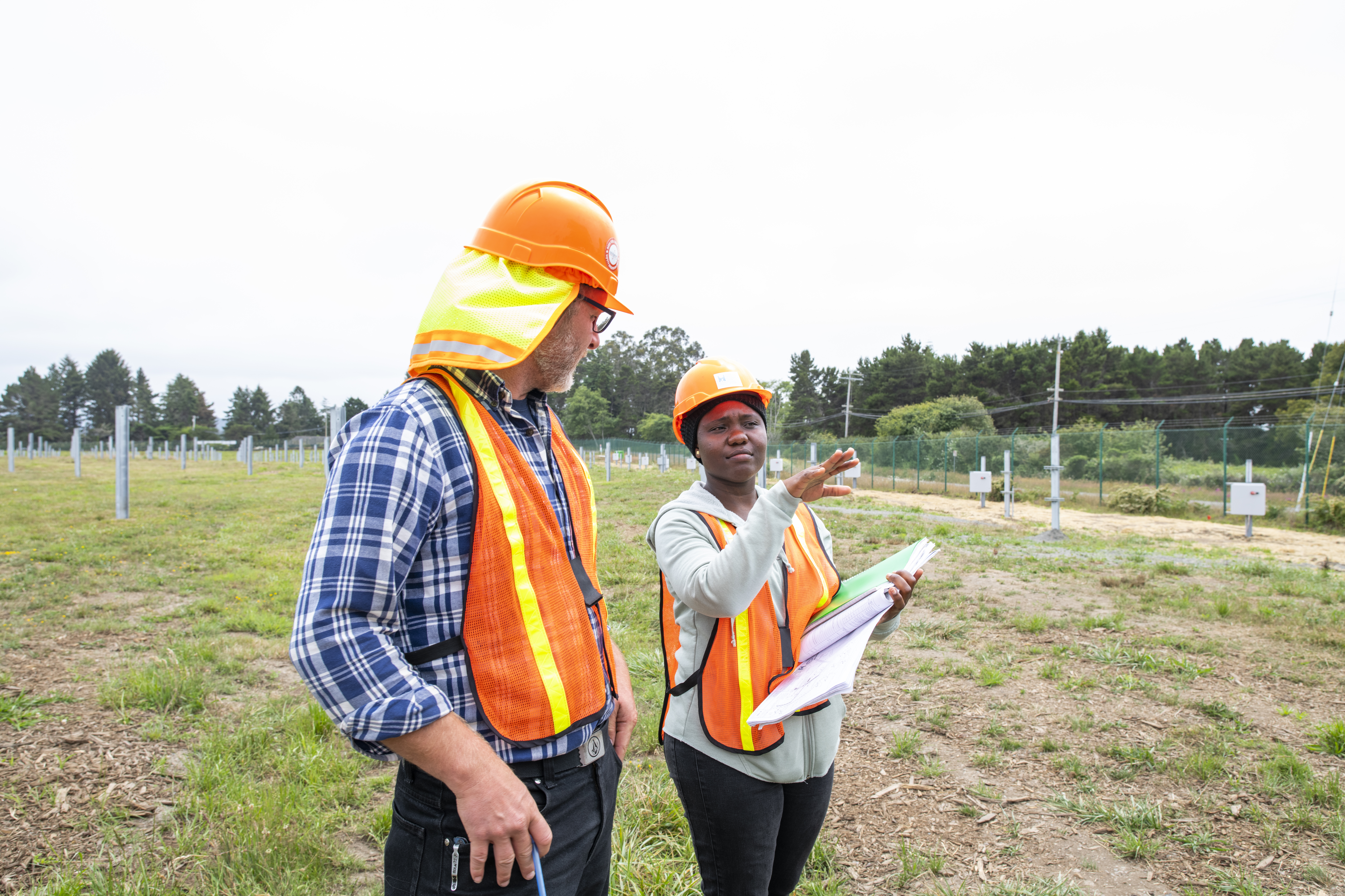 Two engineers in construction gear discuss plans in an open field